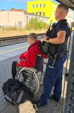 policjant stoi na peronie stacji kolejowej a przed nim na wózku siedzi niepełnosprawny mężczyzna. Obok leży jego torba