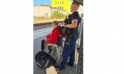 policjant stoi na peronie stacji kolejowej a przed nim na wózku siedzi niepełnosprawny mężczyzna. Obok leży jego torba