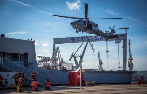 Śmigłowiec Black Hawk zawisa nad statkiem zacumowanym w porcie, na ziemi grupa strażaków.