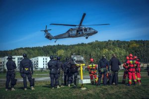 Widok na śmigłowiec Black Hawk w powietrzu na tle błękitnego nieba zza pleców kilkunastu funkcjonariuszy w ciemnych kombinezonach oraz kilku ratowników w pomarańczowych kombinezonach.