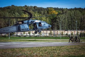 Śmigłowiec Black Hawk podchodzi do lądowania, w tle drzewa. Na betonowym lądowisku na wprost helikoptera znajduje się grupa funkcjonariuszy, z których pierwszy stoi unosząc obie ręce w górę, reszta mężczyzn klęczy.