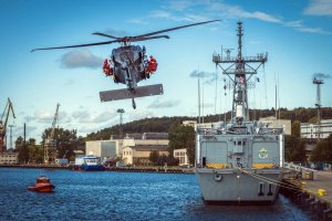 Helikopter Black Hawk zbliża się do zacumowanego w porcie okrętu, po obu stronach śmigłowca zawieszeni są gotowi do desantu funkcjonariusze w pomarańczowych kombinezonach.