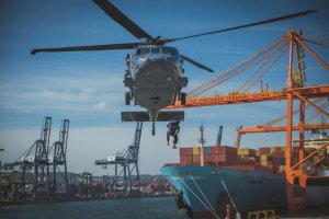 Śmigłowiec Black Hawk znajduje się w zawisie pomiędzy dźwigami portowymi, w tle załadowany kontenerowiec. Dwóch funkcjonariuszy opuszcza się z niego na linach.