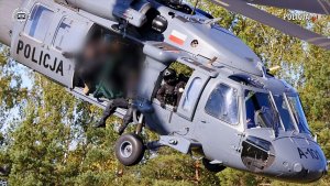 Zbliżenie na bok szarego śmigłowca Black Hawk z duży napisem „POLICJA” – dziób maszyny nachylony jest pod kątem 45 stopni.