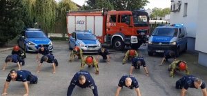 Umundurowani policjanci i strażacy robią pompki na placu, w tle radiowozy