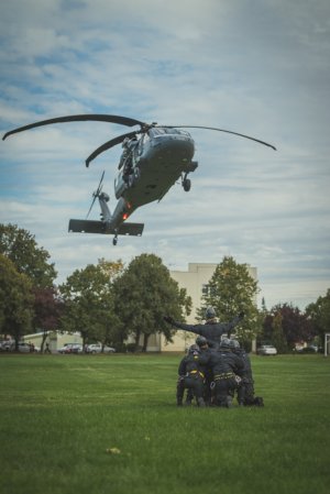 Śmigłowiec Black Hawk unoszący się nad trawiastym lądowisku, w dole grupa ludzi w przyklęku, jeden z nich stoi i daje znaki pilotom.