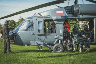 Zbliżenie na przednią część śmigłowca Black Hawk, przed dziobem stoi członek załogi, w otwartych drzwiach przedziału ładunkowego siedzi trzech kontrterrorystów, dwóch z nich trzyma na kolanach grubą linę.