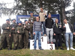 drużyny na podium - Na pierwszym miejscu podium stoi mężczyzna ubrany po cywilnemu, który trzyma w ręku puchar, przed nim stoi jego kolega, na drugim miejscu podium stoi mężczyzna w mundurze polowym z pucharem w ręku ,a obok jego drużyna, na trzecim miejscu podium stoi mężczyzna w ubraniu cywilnym, a obok niego dwaj ubrani po cywilnemu koledzy. Jeden z nich trzyma puchar w ręce.