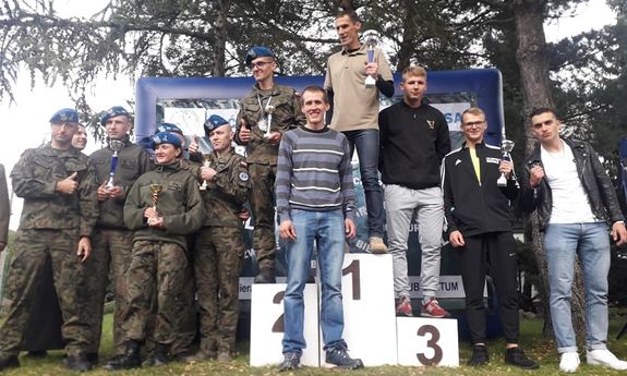 drużyny na podium - Na pierwszym miejscu podium stoi mężczyzna ubrany po cywilnemu, który trzyma w ręku puchar, przed nim stoi jego kolega, na drugim miejscu podium stoi mężczyzna w mundurze polowym z pucharem w ręku ,a obok jego drużyna, na trzecim miejscu podium stoi mężczyzna w ubraniu cywilnym, a obok niego dwaj ubrani po cywilnemu koledzy. Jeden z nich trzyma puchar w ręce.