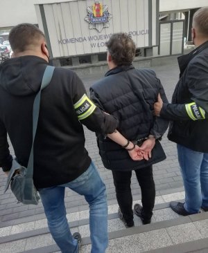 Dziedziniec komendy. Dwóch policjantów po cywilnemu z opaskami na rękawach z napisem Policja, prowadzi zatrzymanego, który ma założone na rękach trzymanych z tyłu kajdanki