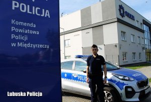 Umundurowany policjant przed budynkiem Komendy Powiatowej Policji w Międzyrzeczu, przed nim z boku widoczny baner z napisem: Policja, Komenda Powiatowa Policji w Międzyrzeczu, za nim widoczny radiowóz