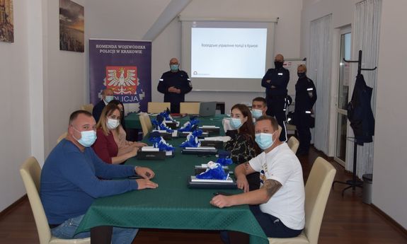 zdjęcie uczestników szkolenia - kilka osób w ubraniach cywilnych i w maseczkach siedzi przy stole, w głębi widocznych jest troje umundurowanych policjantów a za nimi ekran.