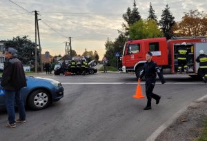 Umundurowany policjant, w tle wóz strażacki i strażacy przy samochodzie osobowym na miejscu wypadku