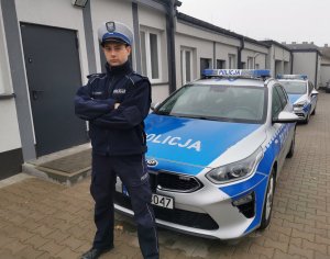 Umundurowany funkcjonariusz stoi przed oznakowanym radiowozem policji