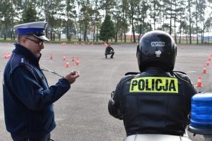 po lewej stronie policjant ruchu drogowego patrzy na stoper, po prawej stronie policjant na motocyklu, w tle plac i tor przeszkód utworzony przez tak zwane pachołki w kolorze pomarańczowo-białym