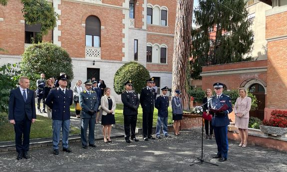 umundurowany generał polskiej Policji stoi przy mikrofonie znajdującym się na statywie. Wokół niego na placyku widać pięciu umundurowanych włoskich funkcjonariuszy, umundurowana polska oficer Policji, trzy ubrane po cywilnemu kobiety i mężczyznę w garniturze. Za tymi osobami widoczna jest grupka ludzi - kobiet i mężczyzn