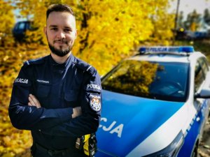 policjant stoi przed radiowozem, w tle drzewa z żółtymi liśćmi