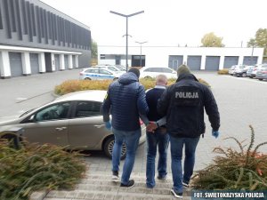 Widok z tyłu. Dwaj policjanci prowadzą mężczyznę. Policjant idący z prawej strony ma na sobie kurtkę z napisem na plecach: Policja. Przed mężczyznami widać parking samochodowy.