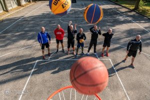 siedmiu mężczyzn stoi na boisku do gry w streetball. Dwóch z nich ma wyciągnięte ręce do góry po rzucie piłki do kosza, jeden z nich trzyma w dłoniach piłkę. Na pierwszym planie widoczne są trzy piłki, które lecą w kierunku kosza