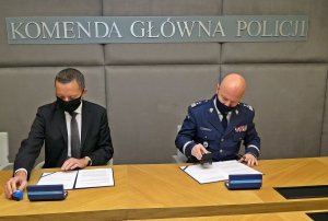 mężczyzna w garniturze i generał Policji siedzą przy stole i podpisują lezące przed nimi dokumenty. Nad mężczyznami widać napis: Komenda Główna Policji