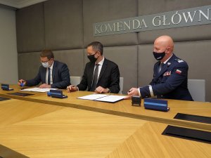 od lewej dwaj mężczyźni w garniturach i generał Policji siedzą przy stole i podpisują leżące przed nimi dokumenty. Nad mężczyznami widać napis: Komenda Główna Policji