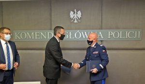 na zdjęciu trzej mężczyźni: od lewej mężczyzna w garniturze, obok drugi mężczyzna w garniturze ściska doń generała Policji. Nad mężczyznami widać napis: Komenda Główna Policji