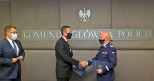 na zdjęciu trzej mężczyźni: od lewej mężczyzna w garniturze, obok drugi mężczyzna w garniturze ściska doń generała Policji. Nad mężczyznami widać napis: Komenda Główna Policji