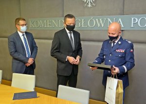 na zdjęciu trzej mężczyźni: od lewej mężczyzna w garniturze, obok drugi mężczyzna w garniturze i generał Policji, który trzyma w dłoniach podarunek i torebkę z logo Poczty Polskiej. Nad mężczyznami widać napis: Komenda Główna Policji