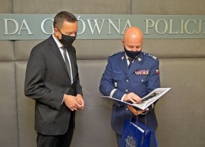 na zdjęciu dwaj mężczyźni: od lewej mężczyzna w garniturze obok generał Policji, który trzyma w dłoniach otwarty album i granatową torebkę z logo Policji. Nad mężczyznami widać napis: Komenda Główna Policji