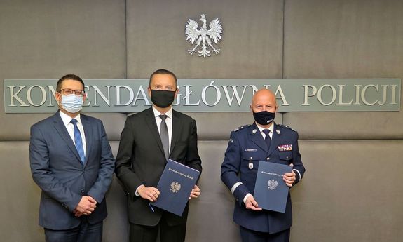 na zdjęciu trzej mężczyźni: od lewej mężczyzna w garniturze, obok drugi mężczyzna w garniturze i generał Policji trzymają w dłoniach granatowe teczki. Nad mężczyznami widać napis: Komenda Główna Policji