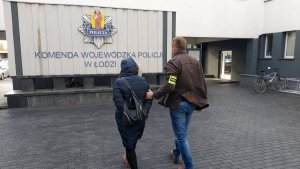 Nieumundurowany policjant prowadzi zatrzymaną. W tle widać budynek a na nim napis: Komenda Wojewódzka Policji w Łodzi