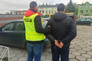 policjant w żółtej kamizelce z napisem Policja trzyma zatrzymanego skutego kajdankami