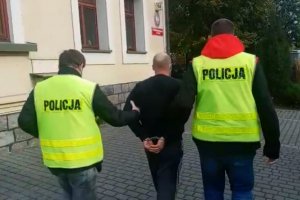 dwaj policjanci w żółtych kamizelkach z napisem Policja prowadzą zatrzymanego skutego kajdankami