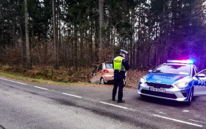 Po prawej stronie radiowóz z włączonymi światłami błyskowymi, po środku policjant drogówki, po lewej pojazd, który zjechał do rowu