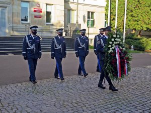 Złożenie wieńca przed obeliskiem &quot;Poległym Policjantom - Rzeczpospolita Polska&quot; na dziedzińcu Komendy Głównej Policji. Na pierwszym planie funkcjonariusz niosący wieniec, w tle komendanci i budynek KGP.