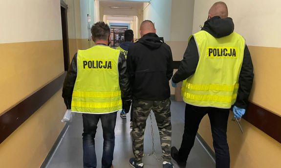 dwaj policjanci w żółtych kamizelkach prowadzą zatrzymanego mężczyznę - widok z tyłu