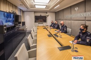 trzej umundurowani oficerowie Policji i policjantka - oficer, siedzą za stołem w sali konferencyjnej. Na przeciwko widoczny jest telewizor i wideokonferencja, za nimi napis: Komenda Główna Policji