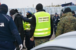 Policjanci wykonują czynności z zatrzymanymi, przy czerwonym busie&quot;&gt;