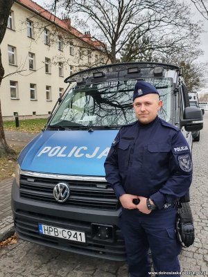 policjant stoi przed radiowozem w tle budynek i drzewa rosnące wzdłuż ulicy na której stoi zaparkowany radiowóz policyjny