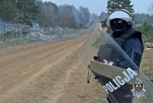 Policjant z kaskiem ochronnym na głowie i tarczą stoi przy drodze gruntowej, w tle graniczne ogrodzenie kolczaste z siatką