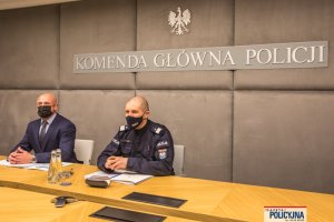 oficer Policji i mężczyzna siedzą w sali za stołem, nad nimi widoczny napis: Komenda Główna Policji