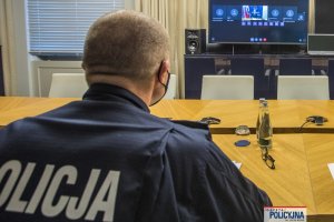 plecy oficera Policji siedzącego za stołem, przed nimi stoi telewizor, na którym widać mężczyznę