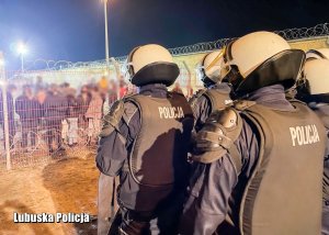 Policjanci oddziałów prewencji Policji stojący przed grupą mężczyzn za siatką ochronną.&quot;&gt;