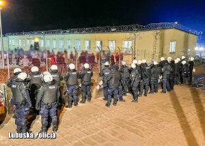 Policjanci oddziałów prewencji Policji stojący przed grupą mężczyzn za siatką ochronną