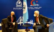 dwaj mężczyźni w maseczkach siedzą w fotelach na przeciwko siebie i rozmawiają. Za nimi w stojakach znajduje się flaga z logo Interpolu, a nad ich głowami na ściance logo Interpolu i napis w języku angielskim Stambuł, Turcja 2021, 89. Zgromadzenie Ogólne