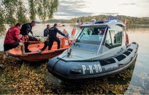 trzej policjanci i ratownik WOPR niosą na noszach osobę przykrytą kocem termicznym. Z boku widać motorową łódź policyjną