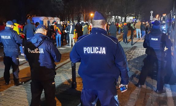 Wieczór. Czterech policjantów stoi przy autobusie na chodniku, a przed nimi widać tłum ludzi - widok z tyłu