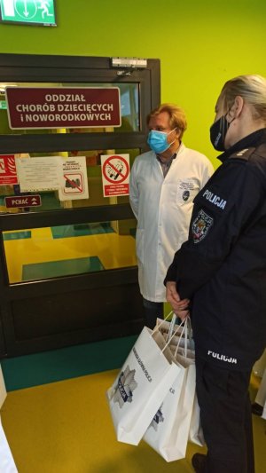 policjantka trzyma w rękach torby z upominkami i wraz z lekarzem stoi przed zamkniętym drzwiami Oddziału Chorób Dziecięcych i Noworodkowych