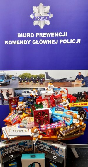 Wypełniony prezentami stolik na tle banneru Biura Prewencji Komendy Głównej Policji. Na stoliku  i pod nim leżą słodycze, pluszowe misie, gry planszowe, słuchawki i tablety. Na banerze widać napis: Policja – Biuro Prewencji Komendy Głównej Policji.