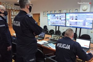 Umundurowani policjanci w pomieszczeniu sztabowym, stoją przodem do ekranów, na których widoczny jest obraz z kamer